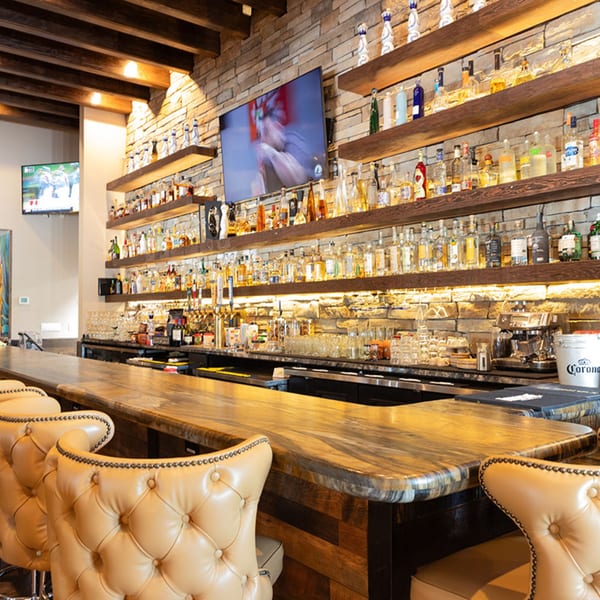 Azteca D'Oro Mexican Restaurant bar with a stone wall, wooden shelves of liquor, and tufted leather bar stools.