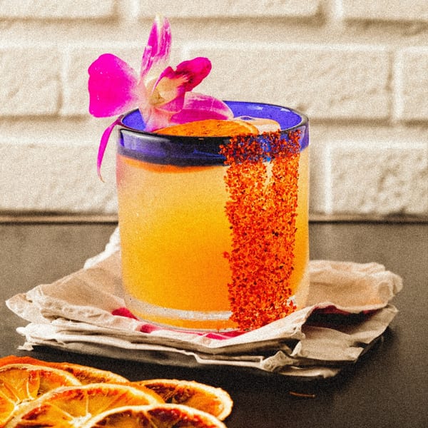 A vibrant orange Mexican cocktail in a blue-rimmed glass, garnished with a pink orchid and chili salt.