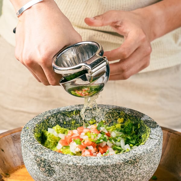 Hands squeezing lime juice into a molcajete filled with guacamole, diced tomatoes, and onions.