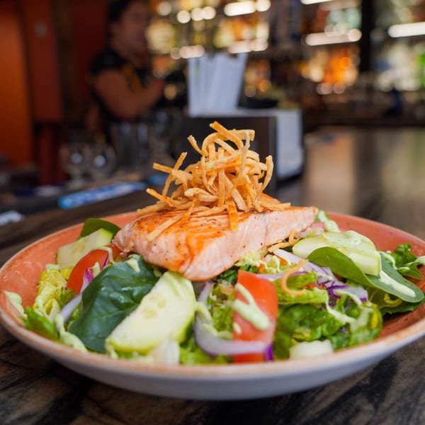 A vibrant salmon salad with mixed greens, tomatoes, cucumbers, and crispy tortilla strips.