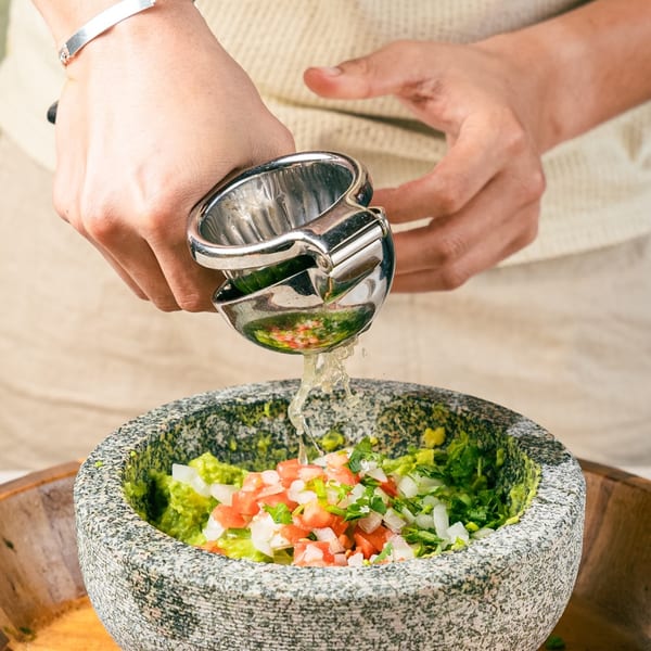 Hands squeezing lime juice into a stone molcajete filled with guacamole, tomatoes, and onions.