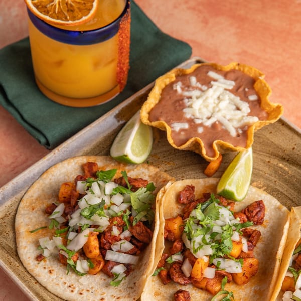 Two al pastor tacos with cilantro and onions, refried beans, and a margarita with a dried orange.