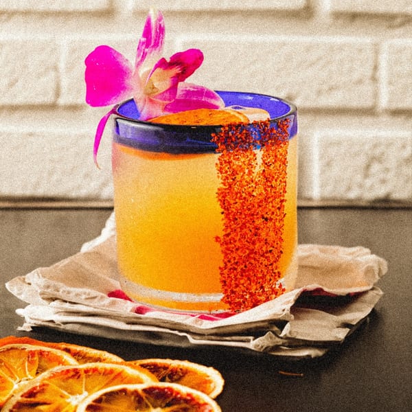 A vibrant orange Mexican cocktail in a blue-rimmed glass, garnished with a pink orchid and chili powder.