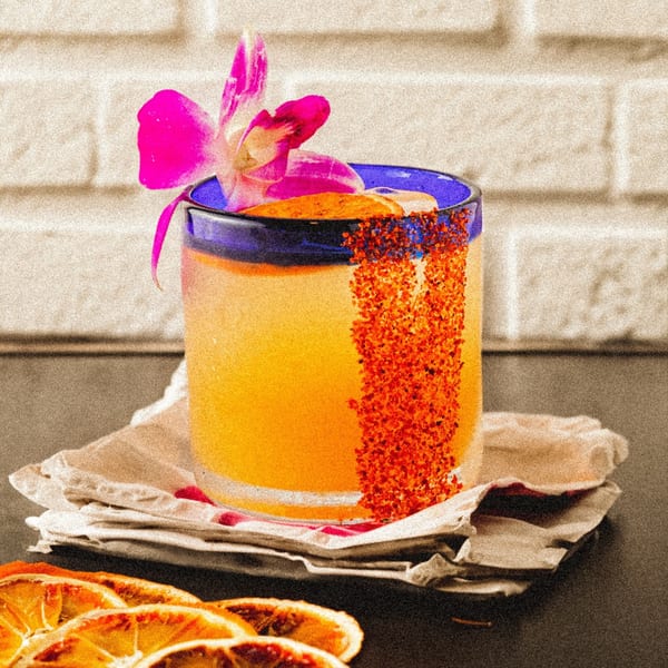 A vibrant orange Mexican cocktail in a blue-rimmed glass, garnished with a pink orchid and chili powder.