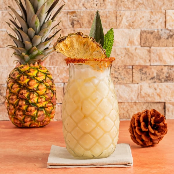 A pineapple-shaped glass of creamy yellow drink with a dried pineapple slice and mint garnish.