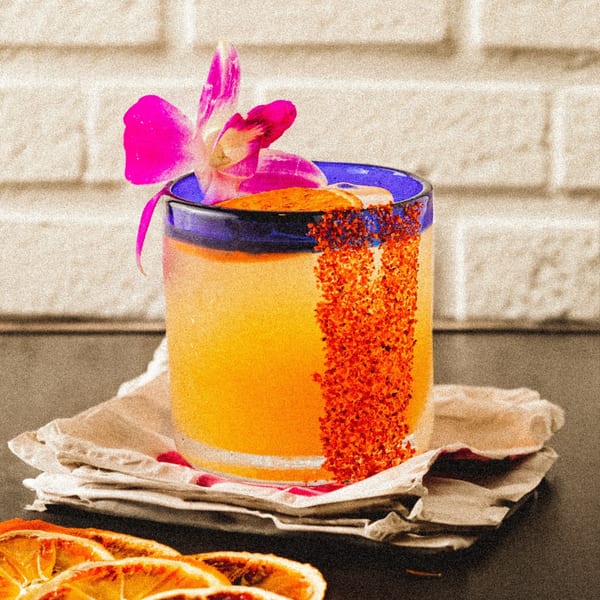 A vibrant orange Mexican cocktail in a blue-rimmed glass, garnished with a pink orchid and chili powder.