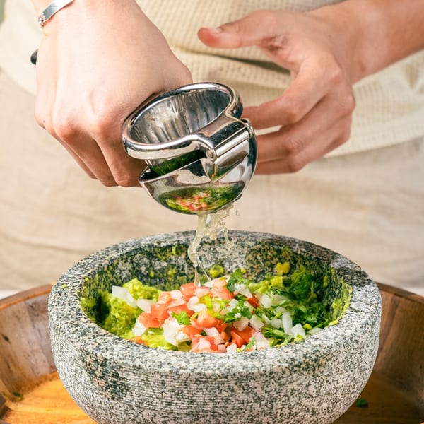 Hands squeezing lime juice into a stone bowl of guacamole with chopped tomatoes and onions.