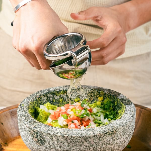 Hands squeezing lime juice into a stone bowl of guacamole with chopped tomatoes and onions.