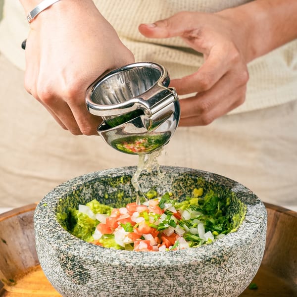 Hands squeezing lime juice into a molcajete filled with guacamole, diced tomatoes, and onions.