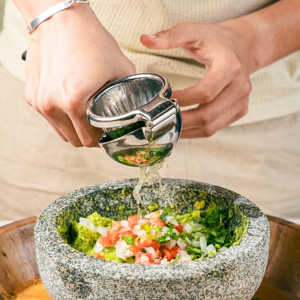 Hands squeezing lime juice into a stone molcajete filled with guacamole, tomatoes, and onions.