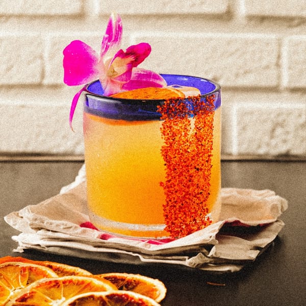 A vibrant orange Mexican cocktail in a blue-rimmed glass, garnished with a pink orchid and chili salt.