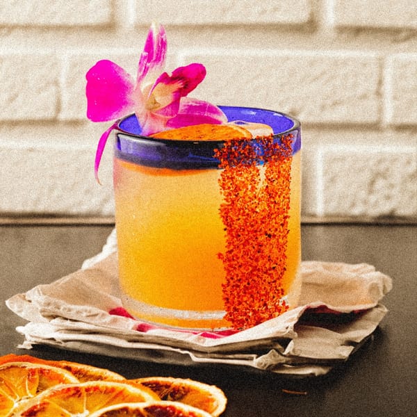 A vibrant Mexican cocktail with a chili-salted rim and a pink orchid garnish, next to dried orange slices.