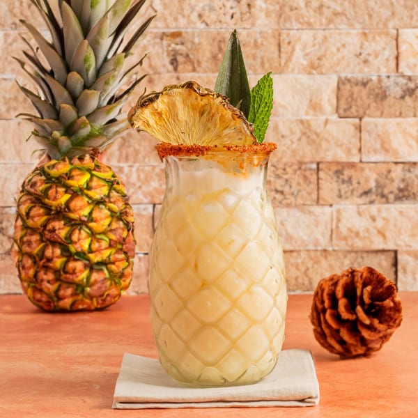 Pineapple-shaped glass filled with a creamy drink, garnished with dried pineapple and mint, beside a whole pineapple.