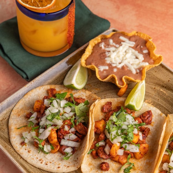 Two tacos with meat, pineapple, onions, and cilantro, with a side of refried beans and a drink.