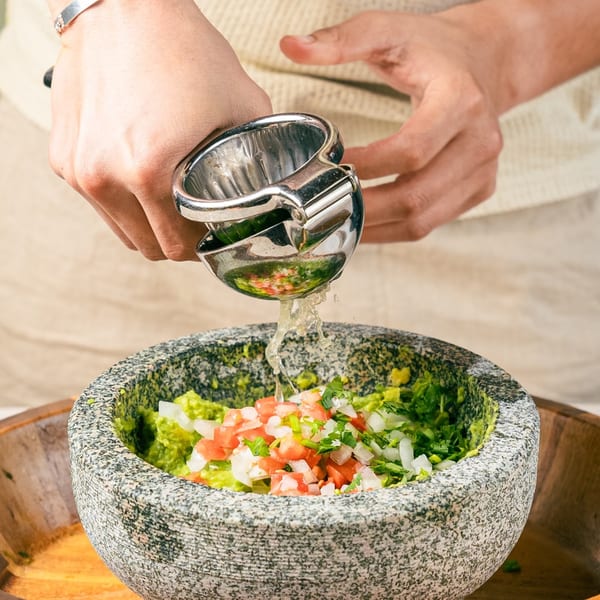 Hands squeezing lime juice into a stone molcajete filled with guacamole, tomatoes, and onions.