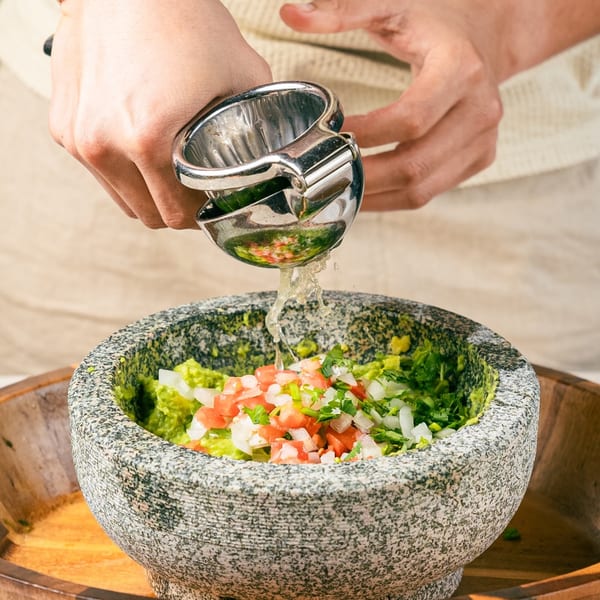 Hands squeezing lime juice into a stone molcajete filled with guacamole, diced tomatoes, and onions.