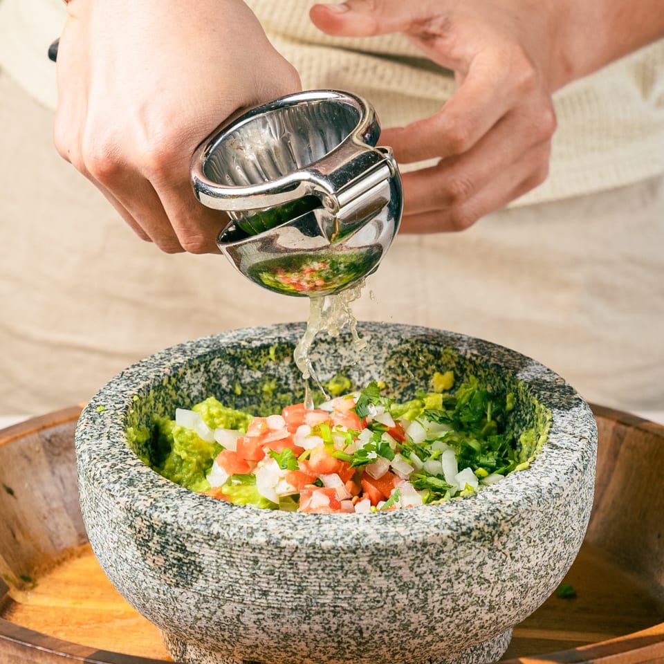 Hands squeezing lime juice into a stone molcajete filled with guacamole, diced tomatoes, and onions.
