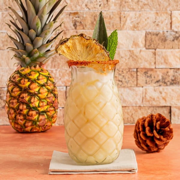 Pineapple-shaped glass filled with a creamy drink, garnished with dried pineapple and mint, next to a whole pineapple.