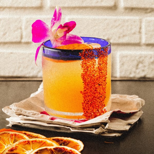 A vibrant orange Mexican cocktail in a blue-rimmed glass, garnished with a pink orchid and chili salt.