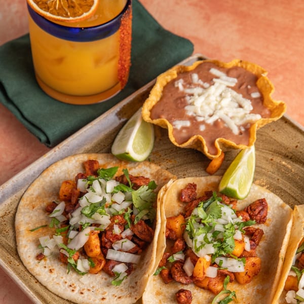 Two al pastor tacos with cilantro and onions, refried beans, and a margarita with an orange slice.