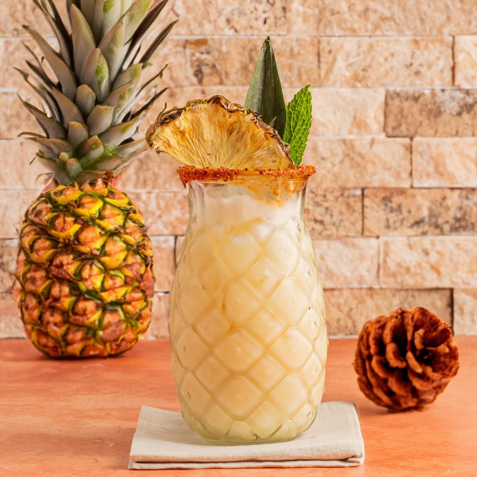 Pineapple drink in a textured glass with a dried pineapple slice and mint, next to a whole pineapple.