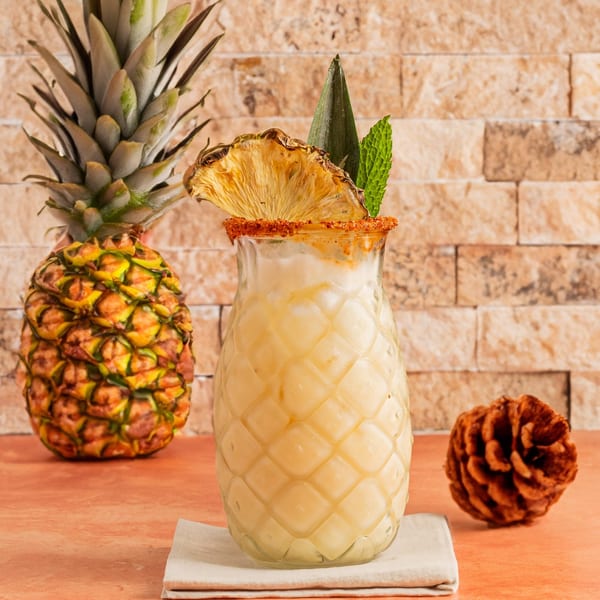 Pineapple-shaped glass filled with a creamy drink, garnished with dried pineapple and mint, next to a whole pineapple.
